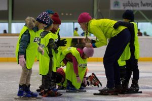 kinderen in een geel hesje staan op schaatsen op het ijs en luisteren naar een coach