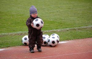 Een peuter in dikke winterkleding bij een voetbalveld houdt een voetbal vast en achter hem liggen er nog 5