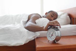 een man ligt in zijn bed en drukt slaperig met 1 hand op een wekker