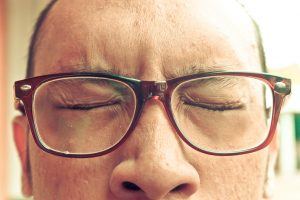 een close up van een gezicht van een man met bril die zijn ogen stevig dichtknijpt