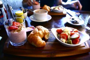 een houten ontbijtplankje met een glaasje aardbeienkwark, een croissantje en een schaaltje fruit