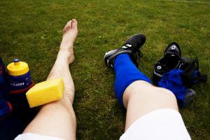 je ziet de benen van een voetballer die een natte spons op zijn knie heeft liggen en 1 sok en schoen uit heeft