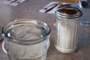 een suikerpotje naast een glas water waar een kopje koffie achter staat