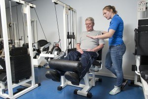 Oudere man doet een krachtoefening voor zijn benen op een fitnessapparaat en luistert naar zijn begeleidster