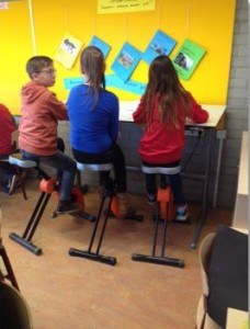 Je ziet 3 kinderen op de rug terwijl ze op een fietsstoel aan een tafel zitten