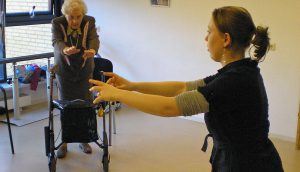 Oudere vrouw met rollator krijgt uitleg over een oefening waarbij ze haar armen voor zich uit moet strekken