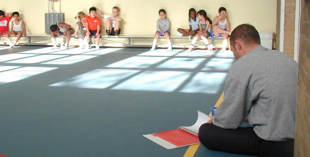 Kinderen zitten op een bankje tijdens de gymles en de gymleraar zit op de grond met een map voor zich