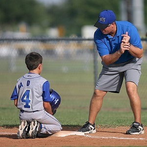 een jongen in honkbalkleding zit op de grond bij een honk en krijgt uitleg van zijn coach