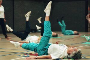 ouderen liggen op hun rug in een sporthal en doen een beenoefening