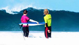 2 surfers op de rug gezien, staan in het water en kijken naar een aankomende golf