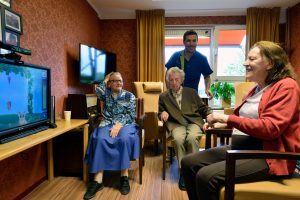 3 ouderen zitten rondom een tv in de huiskamer van een verzorgingshuis