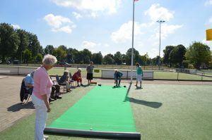 ouderen spelen een balspel waarbij ze een bal naar een lijn op een groene mat moeten rollen buiten op een sportcomplex
