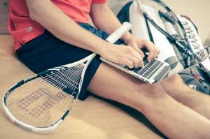 een man zit op de grond van een squashhal met zijn squashraket op zijn schoot en een laptop waarop hij aan het werk is