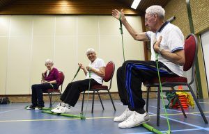 3 ouderen zitten op een stoel en doen oefeningen met een elastiek aan een rekstok