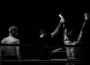 zwart wit foto waarop je 2 boksers op de rug ziet en de scheidsrechter tussen hen in wijst de rechter bokser aan als winnaar