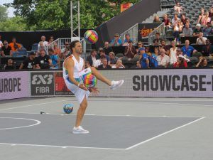 een man jongleert met 2 grote basketballen terwijl publiek op de tribune toekijkt
