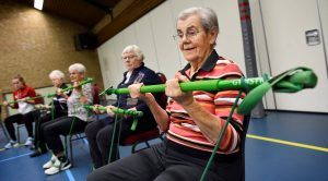 ouderen die zittend op een stoel in een gymzaal een arm oefening doen met een rekstok