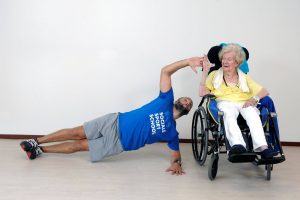 een sportieve man leunt zijdelings op 1 arm op de grond naast een oudere vrouw in een rolstoel
