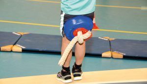 het touw in de gymzaal is omgetoverd als een schommel; kind klemt dit tussen zijn benen en staat op het punt zich af te zetten voor een zwaai naar de andere kant